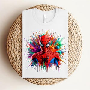 Op de afbeelding: Wit T-shirt met een Spider-Man afbeelding. De superheld is afgebeeld in zijn rood-blauwe pak, tegen een kleurrijke verfspat. Het T-shirt is opgevouwen op een ronde, geweven ondergrond.