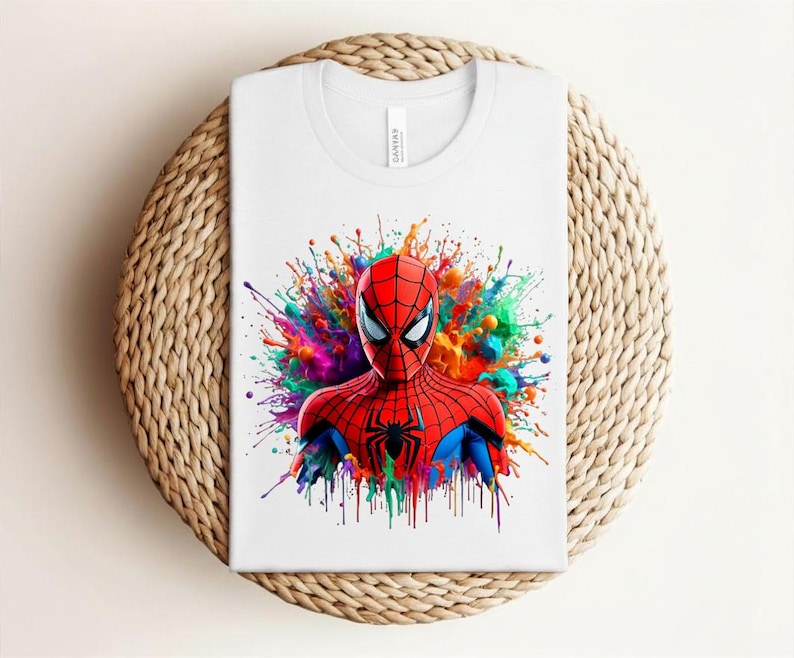 Op de afbeelding: Wit T-shirt met een Spider-Man afbeelding. De superheld is afgebeeld in zijn rood-blauwe pak, tegen een kleurrijke verfspat. Het T-shirt is opgevouwen op een ronde, geweven ondergrond.