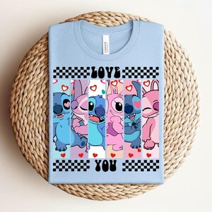 May include: Light blue t-shirt featuring a graphic of cartoon characters in shades of blue and pink. The text "LOVE YOU" is displayed in black and white checkered boxes. Hearts are scattered throughout the design.