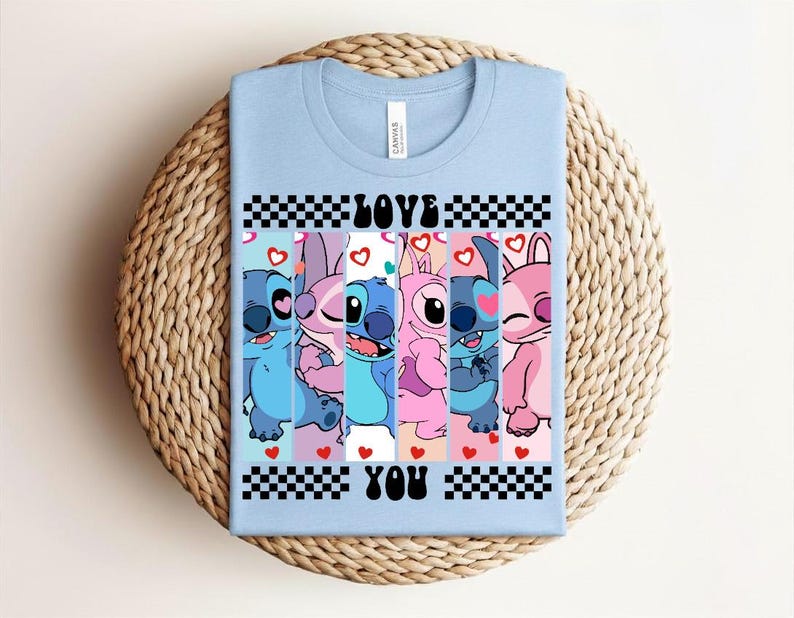 May include: Light blue t-shirt featuring a graphic of cartoon characters in shades of blue and pink. The text "LOVE YOU" is displayed in black and white checkered boxes. Hearts are scattered throughout the design.