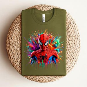 Op de afbeelding: Olijfgroen t-shirt met een kleurrijke Spider-Man-afbeelding. De superheld is afgebeeld in zijn rood-blauwe pak, met een zwart spinnenembleem op zijn borst. De achtergrond heeft levendige verfspatten.