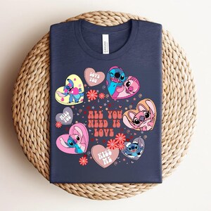Peut inclure: T-shirt bleu marine avec des personnages de dessins anim&eacute;s en forme de c&oelig;ur. Le texte "ALL YOU NEED IS LOVE" est visible, ainsi que d'autres phrases. Le motif comprend des fleurs roses et rouges.