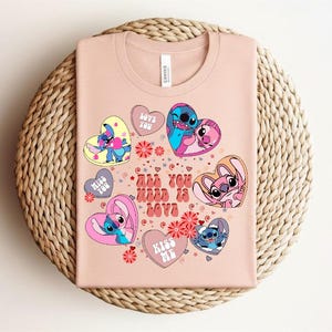 Peut inclure: T-shirt rose clair avec des personnages de dessins anim&eacute;s en forme de c&oelig;ur. Le texte "All You Need Is Love" est affich&eacute;. Le design comprend du bleu, du rose et du jaune. Le t-shirt est pli&eacute; sur un tapis rond tress&eacute;.