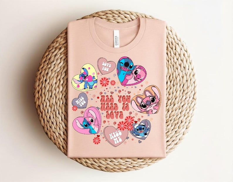 Peut inclure: T-shirt rose clair avec des personnages de dessins anim&eacute;s en forme de c&oelig;ur. Le texte "All You Need Is Love" est affich&eacute;. Le design comprend du bleu, du rose et du jaune. Le t-shirt est pli&eacute; sur un tapis rond tress&eacute;.