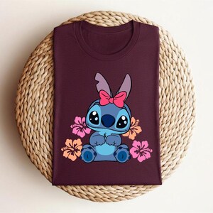 Puede incluir: Camiseta burdeos con un personaje de dibujos animados de color azul, ojos negros grandes y un lazo rosa. Flores de hibisco rosas y naranjas rodean al personaje. La camiseta est&aacute; doblada sobre una superficie circular tejida.