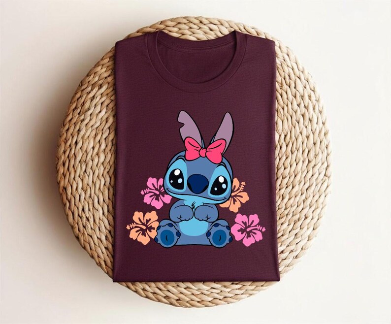 Puede incluir: Camiseta burdeos con un personaje de dibujos animados de color azul, ojos negros grandes y un lazo rosa. Flores de hibisco rosas y naranjas rodean al personaje. La camiseta est&aacute; doblada sobre una superficie circular tejida.
