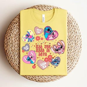 Peut inclure: Un t-shirt jaune avec des personnages de dessins anim&eacute;s dans des motifs en forme de c&oelig;ur. Le t-shirt porte l'inscription "ALL YOU NEED IS LOVE" et d'autres phrases comme "LOVE YOU" et "KISS ME". Le t-shirt est sur une surface tiss&eacute;e.