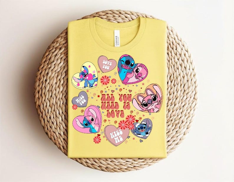 Peut inclure: Un t-shirt jaune avec des personnages de dessins anim&eacute;s dans des motifs en forme de c&oelig;ur. Le t-shirt porte l'inscription "ALL YOU NEED IS LOVE" et d'autres phrases comme "LOVE YOU" et "KISS ME". Le t-shirt est sur une surface tiss&eacute;e.