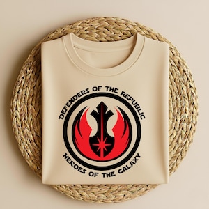 May include: A beige crew neck t-shirt featuring a black and red graphic. The graphic includes a circular design with the text "DEFENDERS OF THE REPUBLIC" and "HEROES OF THE GALAXY". The design also features a stylized emblem with a star.