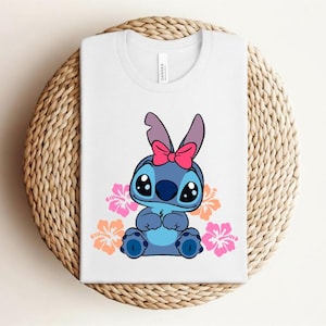 Puede incluir: Camiseta blanca con un personaje de dibujos animados azul, ojos negros grandes y un lazo rosa. Flores de hibisco rosas y naranjas rodean al personaje. La camiseta est&aacute; doblada sobre una estera tejida circular.