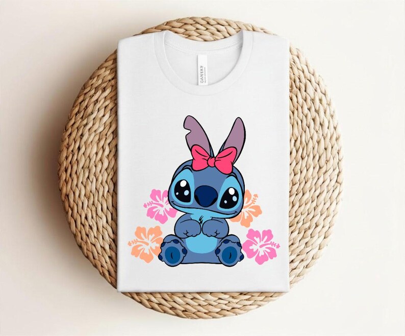 Puede incluir: Camiseta blanca con un personaje de dibujos animados azul, ojos negros grandes y un lazo rosa. Flores de hibisco rosas y naranjas rodean al personaje. La camiseta est&aacute; doblada sobre una estera tejida circular.