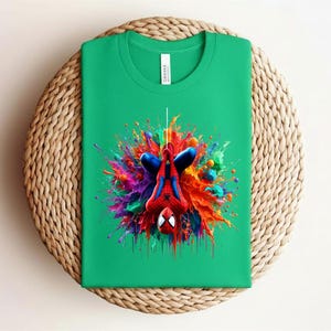 May include: A green t-shirt featuring a colorful graphic of Spider-Man hanging upside down. The design includes vibrant splashes of red, blue, yellow, and purple. The shirt is folded and placed on a woven, circular surface.