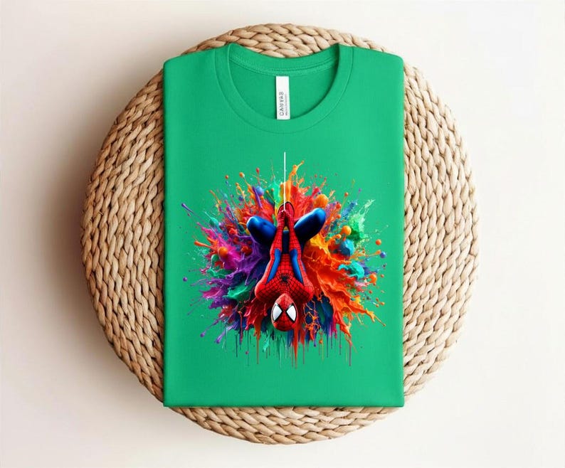 May include: A green t-shirt featuring a colorful graphic of Spider-Man hanging upside down. The design includes vibrant splashes of red, blue, yellow, and purple. The shirt is folded and placed on a woven, circular surface.