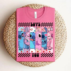 May include: A pink t-shirt featuring a graphic of several cartoon characters with the words "LOVE YOU" in black. The characters are in various shades of blue and pink, with hearts and checkered patterns.