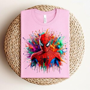 Op de afbeelding: Een lichtroze t-shirt met een kleurrijke afbeelding van Spider-Man. Het gezicht en bovenlichaam van de superheld zijn gedetailleerd afgebeeld, met een levendige splash van kleuren erachter. Het shirt is opgevouwen op een geweven, cirkelvormig oppervlak.