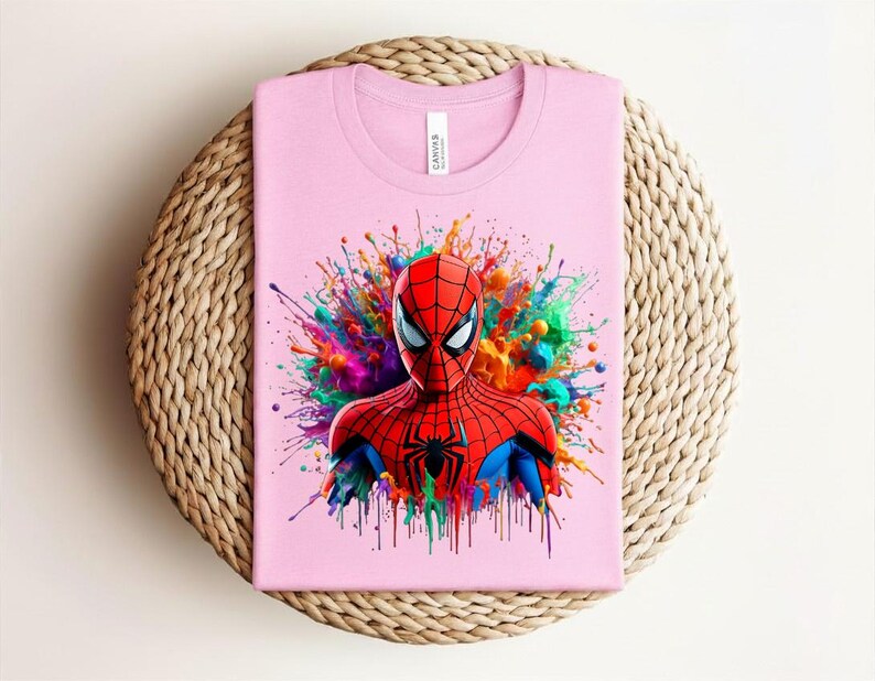 Op de afbeelding: Een lichtroze t-shirt met een kleurrijke afbeelding van Spider-Man. Het gezicht en bovenlichaam van de superheld zijn gedetailleerd afgebeeld, met een levendige splash van kleuren erachter. Het shirt is opgevouwen op een geweven, cirkelvormig oppervlak.
