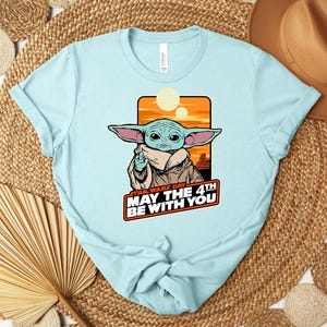 Może przedstawiać: Jasnoniebieska koszulka z grafiką Grogu z Gwiezdnych Wojen, z napisem "Star Wars Day May the 4th Be With You". Projekt zawiera pomarańczowe i ż&oacute;łte tło. Koszulka jest wykonana z miękkiego materiału.