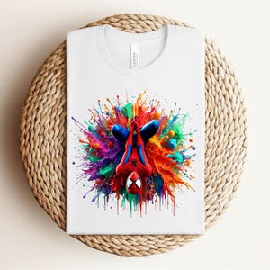 May include: White t-shirt featuring a colorful graphic of Spider-Man in a dynamic pose, surrounded by a vibrant splash of paint in various colors. The design is centered on the shirt.