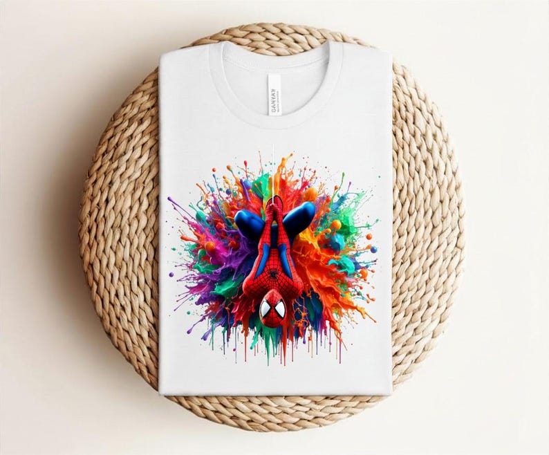 May include: White t-shirt featuring a colorful graphic of Spider-Man in a dynamic pose, surrounded by a vibrant splash of paint in various colors. The design is centered on the shirt.