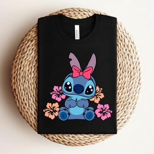 Puede incluir: Camiseta negra con un personaje de dibujos animados azul, un lazo rosa y ojos grandes, rodeado de flores. La camiseta est&aacute; doblada sobre una superficie tejida. Prenda de vestir informal.