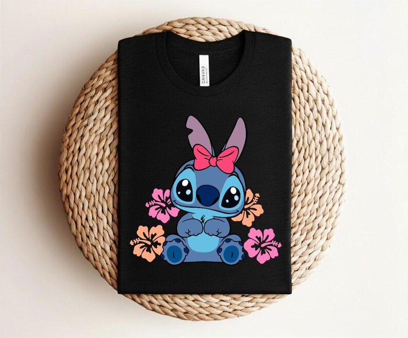 Puede incluir: Camiseta negra con un personaje de dibujos animados azul, un lazo rosa y ojos grandes, rodeado de flores. La camiseta est&aacute; doblada sobre una superficie tejida. Prenda de vestir informal.