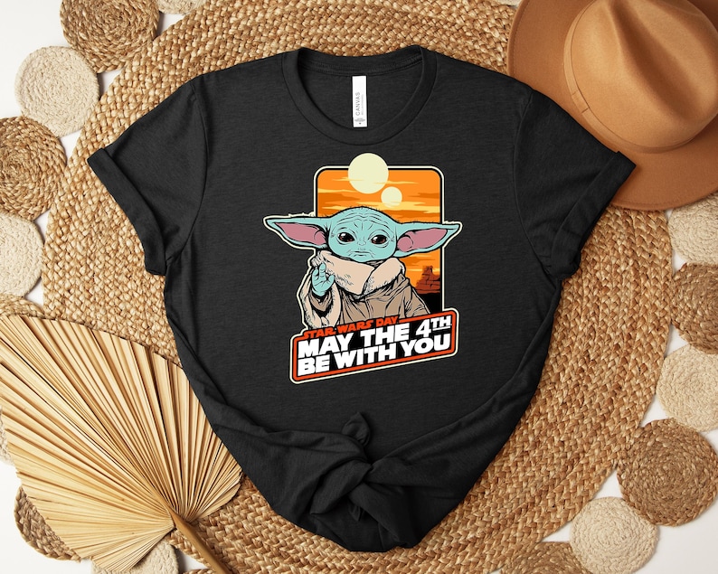 Może przedstawiać: Czarna koszulka z grafiką zielonego kosmity z dużymi uszami na pomarańczowo-ż&oacute;łtym tle. Tekst "STAR WARS DAY MAY THE 4TH BE WITH YOU" jest wyświetlany pod postacią.