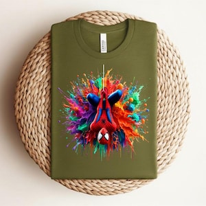 May include: Olive green t-shirt featuring a colorful graphic of Spider-Man hanging upside down. The superhero is surrounded by a vibrant splash of paint in shades of red, blue, yellow, and purple. The shirt is folded on a woven surface.