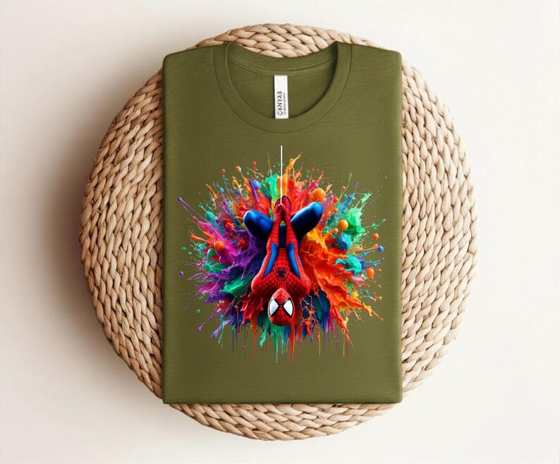May include: Olive green t-shirt featuring a colorful graphic of Spider-Man hanging upside down. The superhero is surrounded by a vibrant splash of paint in shades of red, blue, yellow, and purple. The shirt is folded on a woven surface.