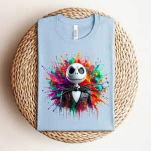 May include: A light blue t-shirt featuring a graphic of a white skull character with a stitched mouth wearing a black suit, centered against a vibrant, multi-colored paint splatter design with hues of red, orange, yellow, green, and purple.