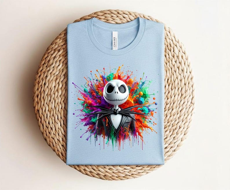 May include: A light blue t-shirt featuring a graphic of a white skull character with a stitched mouth wearing a black suit, centered against a vibrant, multi-colored paint splatter design with hues of red, orange, yellow, green, and purple.