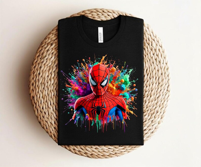 Op de afbeelding: Zwart t-shirt met een kleurrijke afbeelding van Spider-Man. De superheld is afgebeeld in zijn klassieke rood-blauwe pak, met een levendige splash van kleuren erachter. Het shirt is op een geweven oppervlak gevouwen.