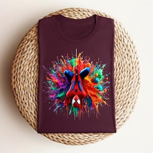May include: A maroon t-shirt featuring a colorful graphic of Spider-Man hanging upside down. The superhero is surrounded by a vibrant explosion of paint in shades of blue, red, green, and yellow. The shirt is displayed on a woven, circular surface.