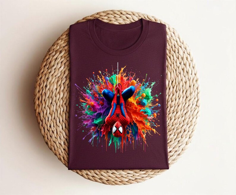 May include: A maroon t-shirt featuring a colorful graphic of Spider-Man hanging upside down. The superhero is surrounded by a vibrant explosion of paint in shades of blue, red, green, and yellow. The shirt is displayed on a woven, circular surface.