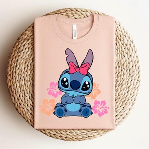 Puede incluir: Camiseta rosa claro con un personaje de dibujos animados azul, adornado con un lazo rosa. El dise&ntilde;o incluye flores de hibisco rosas y naranjas. La camiseta est&aacute; doblada sobre una estera redonda de mimbre.