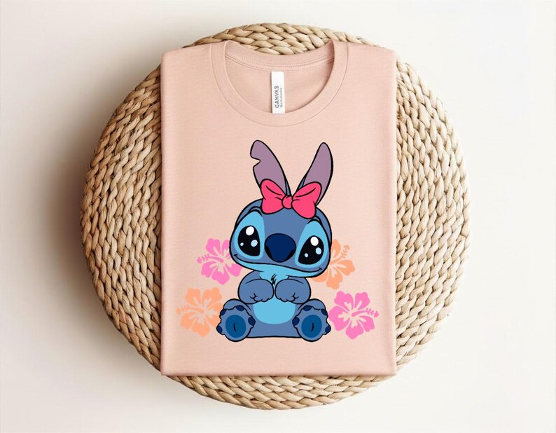 Puede incluir: Camiseta rosa claro con un personaje de dibujos animados azul, adornado con un lazo rosa. El dise&ntilde;o incluye flores de hibisco rosas y naranjas. La camiseta est&aacute; doblada sobre una estera redonda de mimbre.