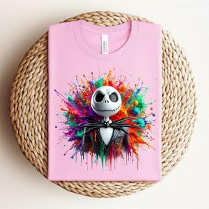 May include: A light pink t-shirt featuring a graphic of a white skull character with large black eyes and a stitched mouth, wearing a black pinstripe suit and bow tie, set against a vibrant, multi-colored paint splatter background.
