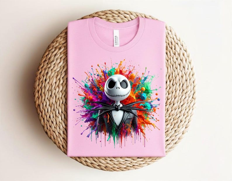 May include: A light pink t-shirt featuring a graphic of a white skull character with large black eyes and a stitched mouth, wearing a black pinstripe suit and bow tie, set against a vibrant, multi-colored paint splatter background.