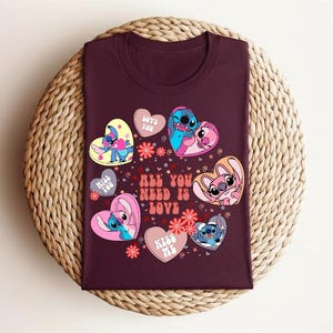 Peut inclure: Un t-shirt bordeaux avec un motif graphique color&eacute; repr&eacute;sentant des personnages de dessins anim&eacute;s en forme de c&oelig;ur. Le texte "ALL YOU NEED IS LOVE" est &eacute;galement inclus. Le t-shirt est pr&eacute;sent&eacute; sur une surface circulaire tiss&eacute;e.