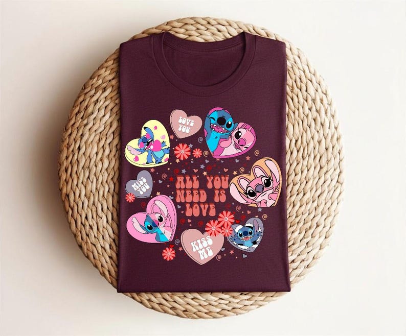 Peut inclure: Un t-shirt bordeaux avec un motif graphique color&eacute; repr&eacute;sentant des personnages de dessins anim&eacute;s en forme de c&oelig;ur. Le texte "ALL YOU NEED IS LOVE" est &eacute;galement inclus. Le t-shirt est pr&eacute;sent&eacute; sur une surface circulaire tiss&eacute;e.