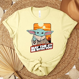 Może przedstawiać: Jasnoż&oacute;łta koszulka z grafiką Grogu z Gwiezdnych Wojen. Obraz zawiera tekst "Star Wars Day May the 4th Be With You" w czerwonej i białej czcionce. Projekt jest osadzony na tle pomarańczowo-ż&oacute;łtego zachodu słońca.