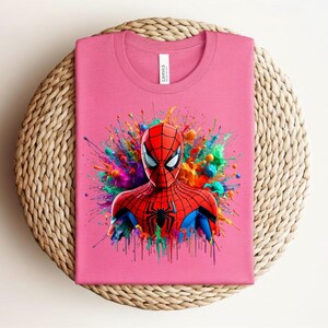 Op de afbeelding: Roze t-shirt met een kleurrijke Spider-Man afbeelding. De superheld is afgebeeld in een rood en blauw pak met een zwart spinnenembleem, tegen een achtergrond van levendige kleurexplosies. Het t-shirt is op een geweven oppervlak gevouwen.