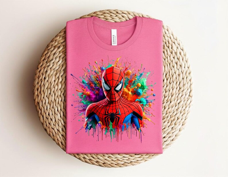Op de afbeelding: Roze t-shirt met een kleurrijke Spider-Man afbeelding. De superheld is afgebeeld in een rood en blauw pak met een zwart spinnenembleem, tegen een achtergrond van levendige kleurexplosies. Het t-shirt is op een geweven oppervlak gevouwen.
