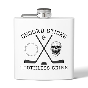 May include: White flask with a silver cap and hinge. The front features a black and white graphic with crossed hockey sticks, a skull, and the text "CROOKD STICKS & TOOTHLESS GRINS". The flask is designed for carrying liquids.