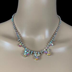 May include: A silver-toned necklace featuring a continuous strand of round, iridescent, aurora borealis-style crystals. The center of the necklace displays a cluster of three dangling crystal drops, each composed of multiple round stones.