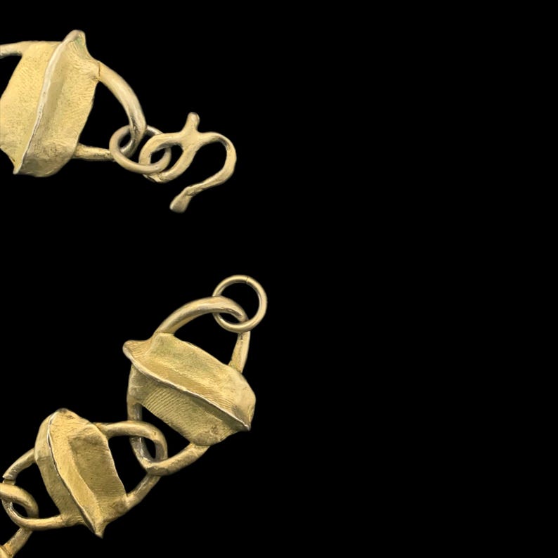 May include: A gold-toned metal chain necklace featuring a series of textured, leaf-shaped links connected by circular rings. The necklace includes a hook-style clasp mechanism at one end, showcasing a matte finish on the metallic surface.