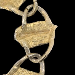 May include: A gold-toned metal chain bracelet featuring textured, rectangular links connected by smooth, rounded loops. A small rectangular plaque is centered on one of the textured links, clearly engraved with the brand name BAUPLAST.