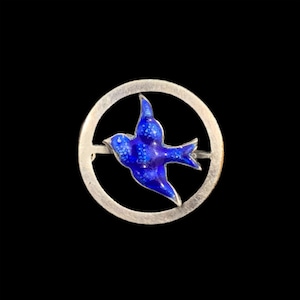 May include: A circular silver-toned brooch featuring a central blue enamel bird in flight. The bird has a deep blue color with lighter blue dot patterns on its wings and body. The bird is positioned within a plain, smooth silver-toned circular frame.
