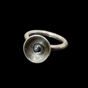May include: A silver ring featuring a bowl-shaped setting with a textured interior surface. A small, dark, spherical bead is centred within the concave bowl, secured by a tiny silver pin. The band of the ring is smooth, polished, and rounded.