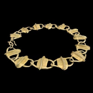 May include: A gold-toned metal chain necklace composed of repeating leaf-shaped links. Each link features a textured surface with a central raised ridge, connected by small circular rings. The necklace includes a simple hook-and-eye clasp closure.