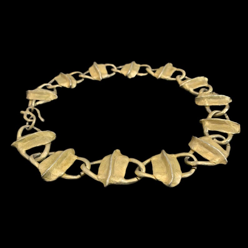 May include: A gold-toned metal chain necklace composed of repeating leaf-shaped links. Each link features a textured surface with a central raised ridge, connected by small circular rings. The necklace includes a simple hook-and-eye clasp closure.